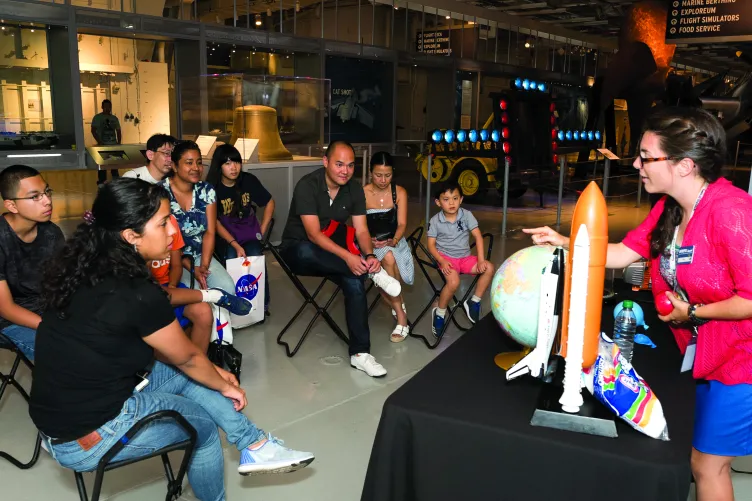 A Museum educator is standing behind a table that has a globe and a model rocket on it and is speaking to a group of seated visitors.