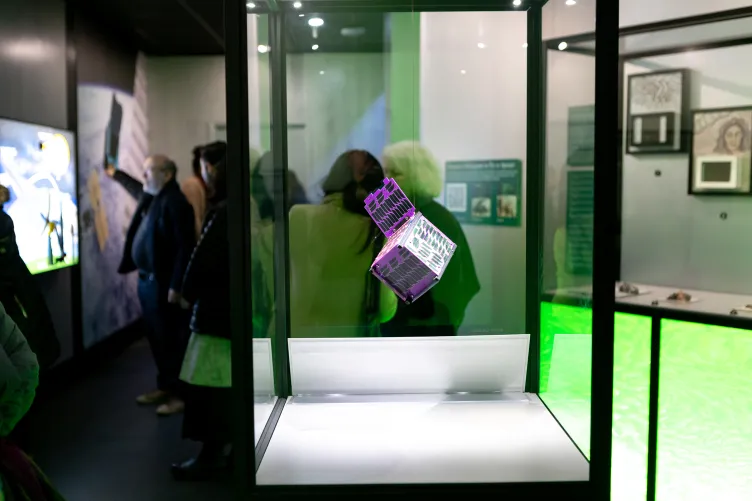 A holographic cube is suspended in mid-air in a display case while visitors are exploring the exhibition behind it.