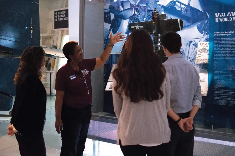 A tour guide is pointing at something in a display case while visitors attentively listen.