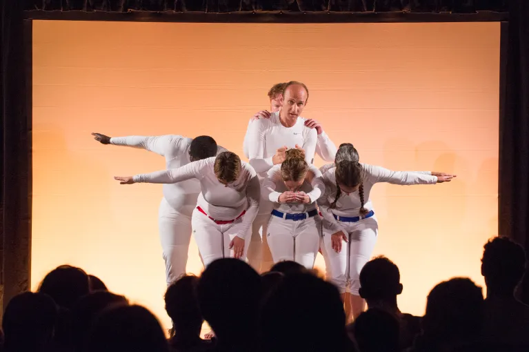 A group of six performers on stage, dressed in white against an orange background, during a performance of Moon Shot.