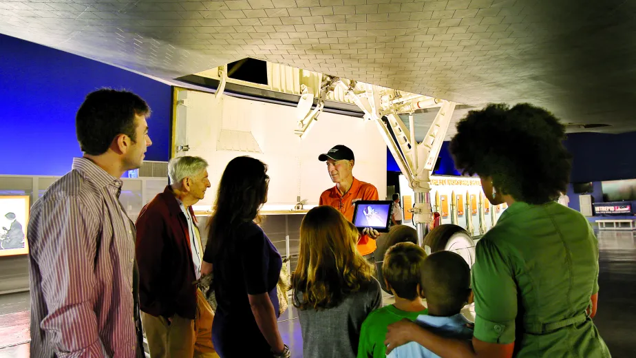 A group of adults and children listening to a tour guide under the Space Shuttle Enterprise.