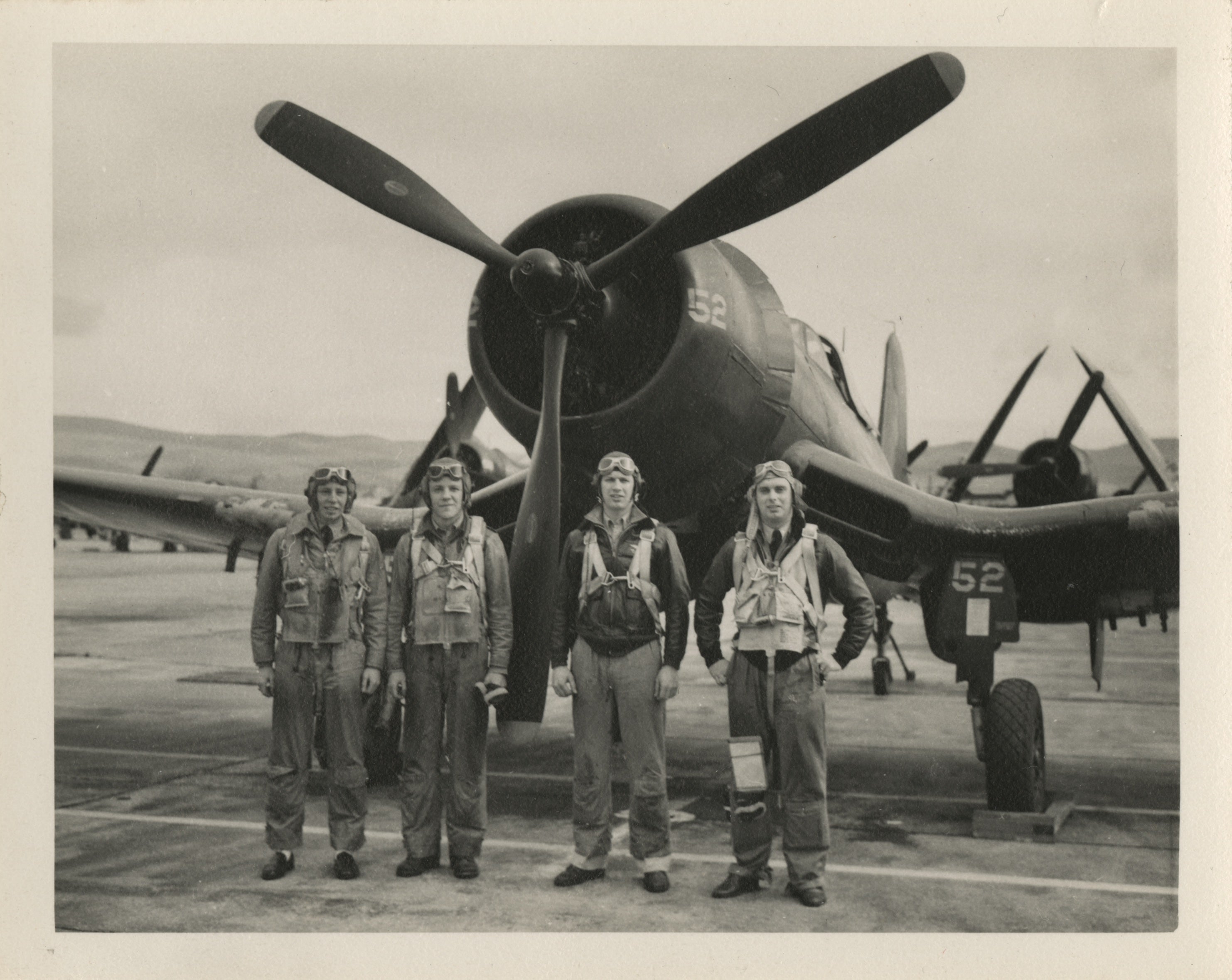 An archival photo of four former crew members standing in front of an FG-1D Corsair on an aircraft carrier flight deck.