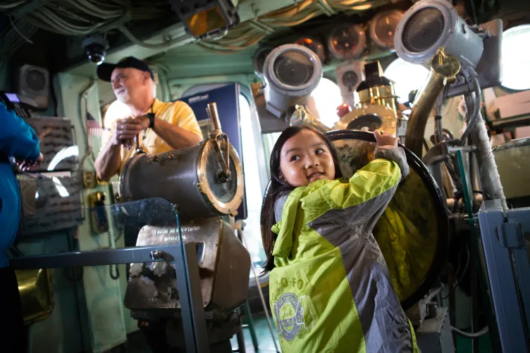 A young girl plays in the ship&#039;s island, spinning the ship&#039;s wheel as a former Intrepid crew member looks on