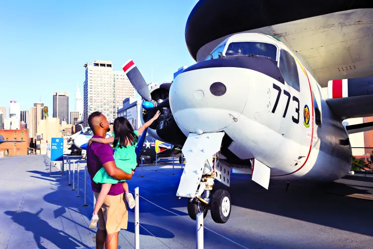 A man and woman read an exhibit panel on Intrepid's flight deck while their child points at the propeller of an aircraft.