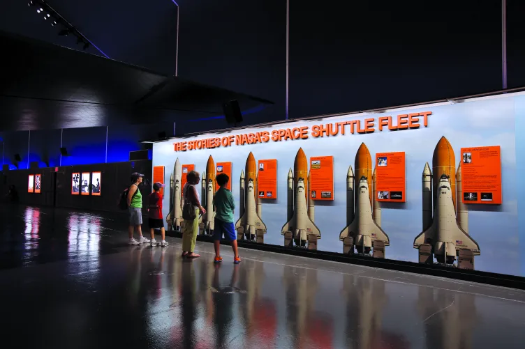 A group of visitors is standing in front of an exhibition panel that reads: "The Stories of NASA's Space Shuttle Fleet."