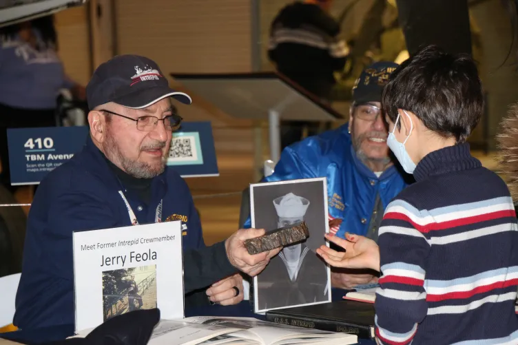 A veteran is seated at a table and showing an artifact to a child.