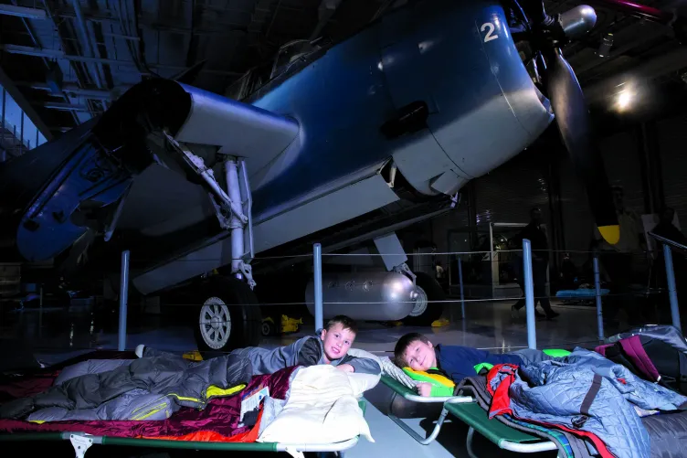 Two kids in sleeping bags are laying on top of cots on the hangar deck next to an airplane