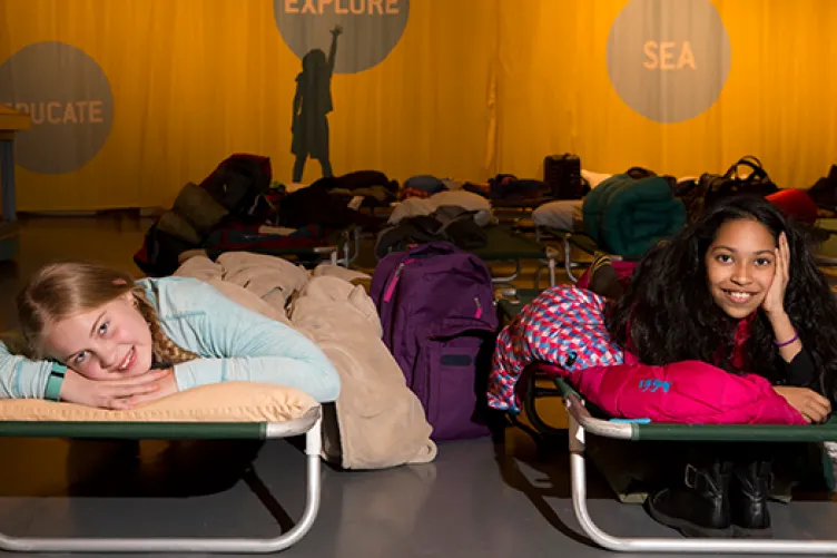 Two kids in sleeping bags are laying on top of cots on the hangar deck