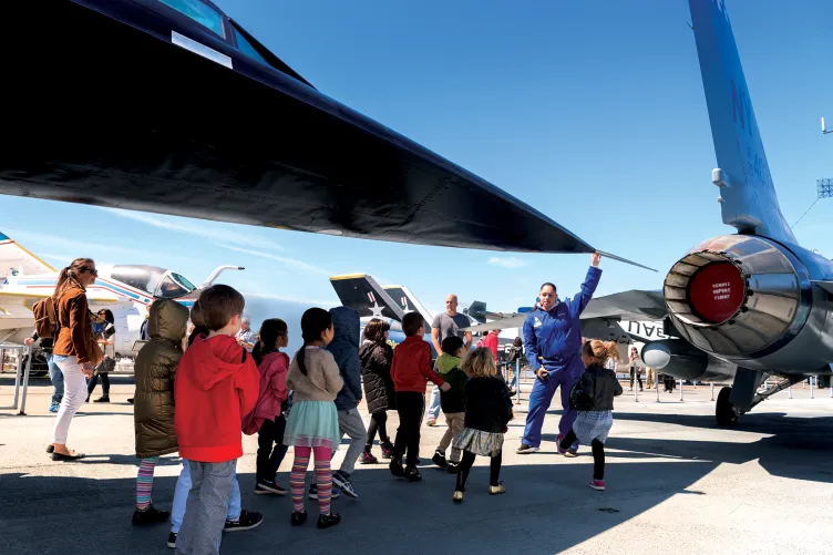 A group of kids at a birthday party are with a Museum educator and looking under the wing of an airplane.
