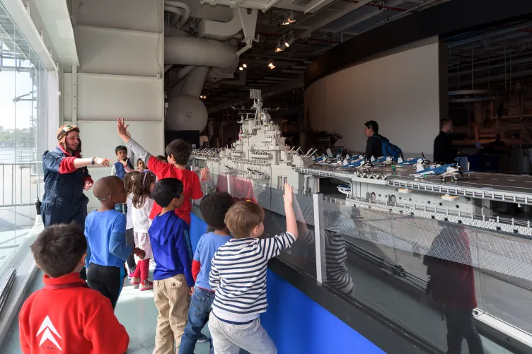 A group of kids are looking at the LEGO model of the Intrepid with a Museum educator who&#039;s wearing a pilot&#039;s helmet.