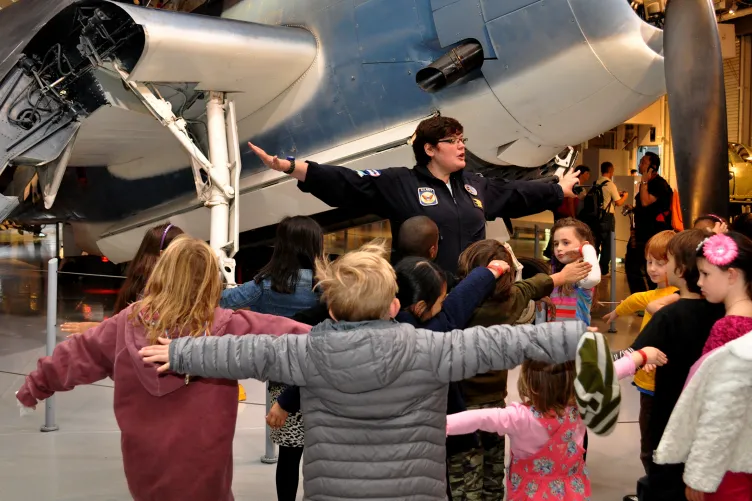 A group of kids at a birthday party are on the hangar deck with their arms outstretched like an airplane.