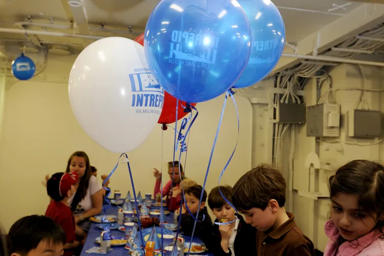 Kids are eating at a table decorated with a blue tablecloth and balloons.