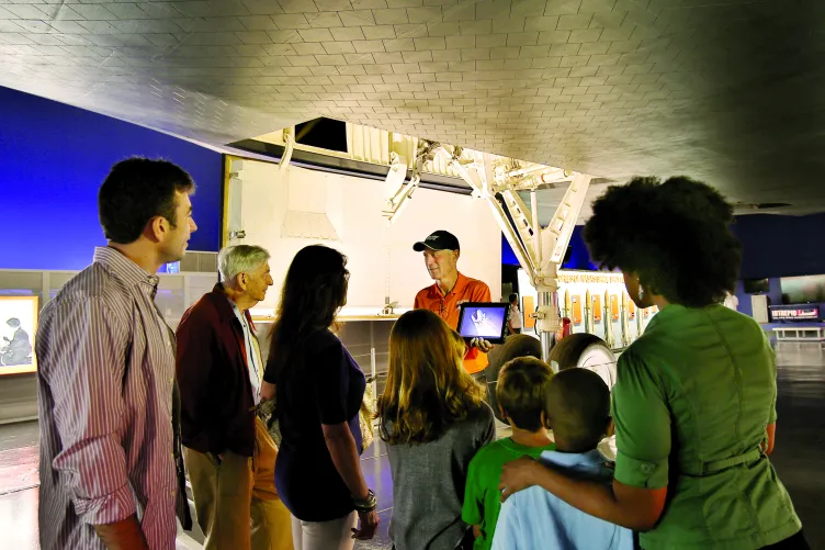 A group of adults and children listening to a tour guide under the Space Shuttle Enterprise.