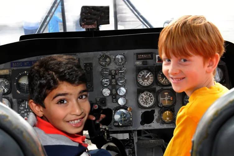 Two kids are seated in the cockpit of an airplane and looking back at the camera smiling.