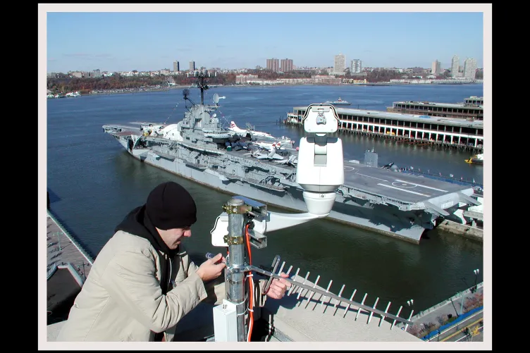 A photo of a person installing a live webcam that was used to document the rebuilding of Pier 86, with the Intrepid in the background.