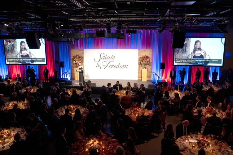 Intrepid Museum president Susan Marenoff-Zausner is speaking at a podium at the Museum's gala event with guests at tables seated nearby.