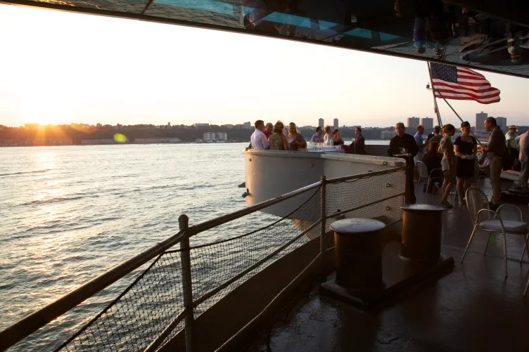 Guests are mingling at an event on the fantail as the sun is setting over the Hudson River. An American flag hangs with them.