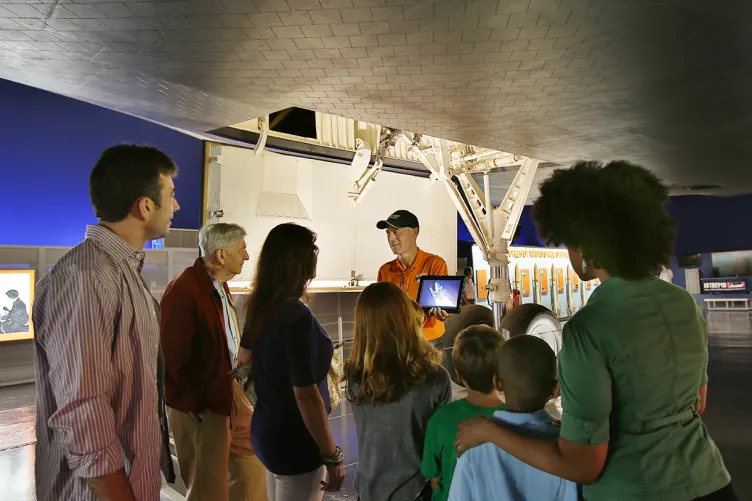 A tour guide is under the Enterprise while speaking to a group and showing them an image on a tablet.