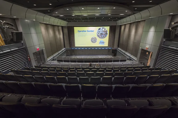A stage set with panelist chairs and a podium in front of a large screen, with theater-style rows of seating in front of them