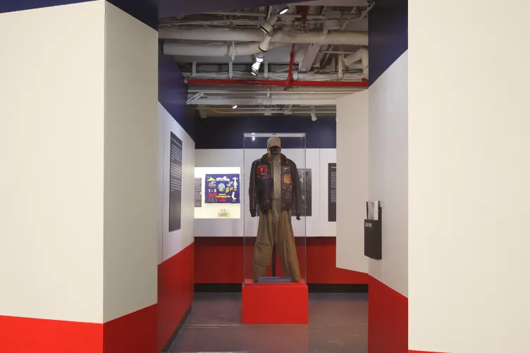 An opening to an exhibition that says “Thirty Years of Collecting: Treasures from Intrepid’s Collections” and an encased uniform in the background.