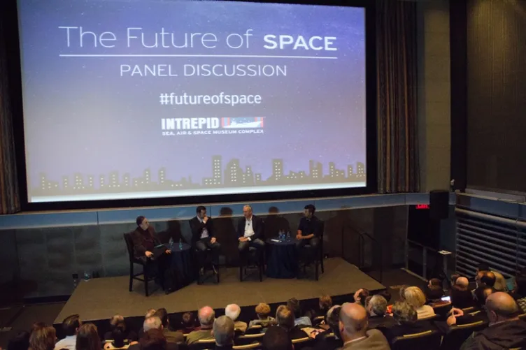 Panelists are seated on a stage front of a large screen, with theater-style rows of people seated in front of them