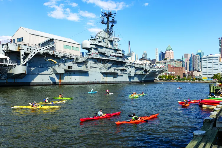 Kayakers are in the Hudson River with the Intrepid in the background.