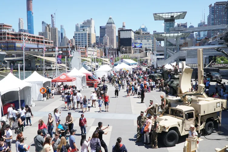 Crowds of people are on Pier 86, with white event tents on the left, and active duty military personnel are sitting on military vehicles on the right.