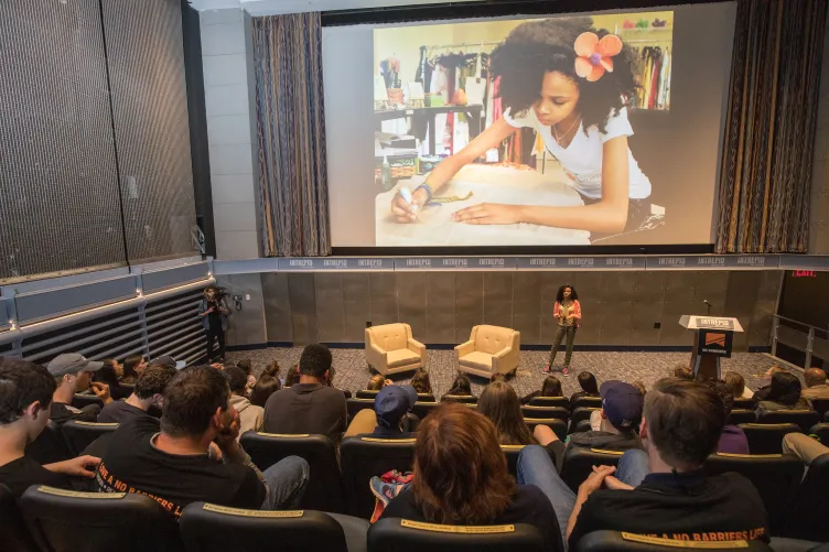 People seated in a theater with someone at the stage and a photo of a child doing an activity on a large screen