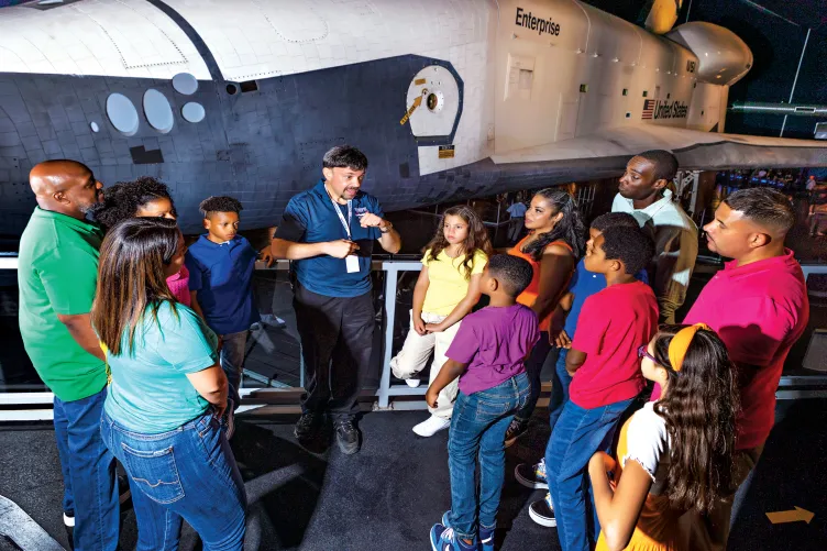 A group of visitors stand in front of the Space Shuttle Enterprise and are listening to a tour guide.