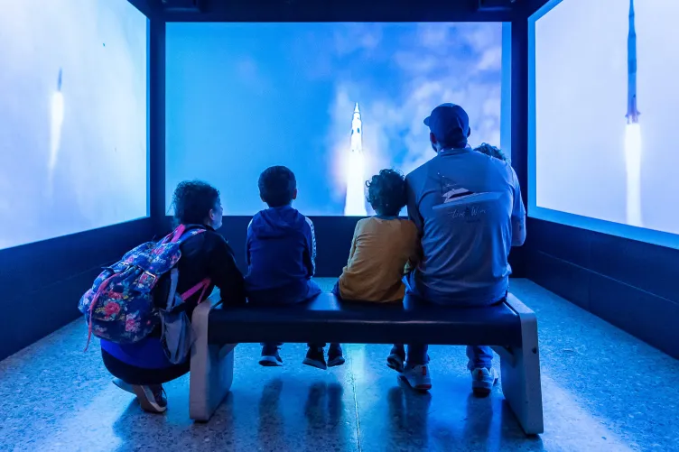 Visitors seated on a bench surrounded by large screens displaying a Saturn V liftoff immersive experience