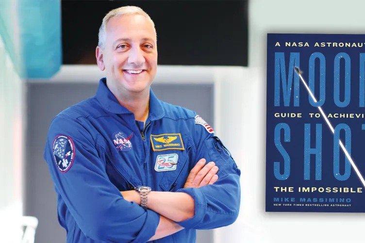Former NASA Astronaut Mike Massimino in a space jumpsuit standing next to his new book titled 'MOONSHOT: A NASA Astronaut’s Guide to Achieving the Impossible.