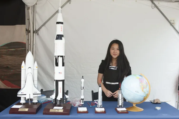A girl is standing at a table, examining replicas of rockets and a globe.