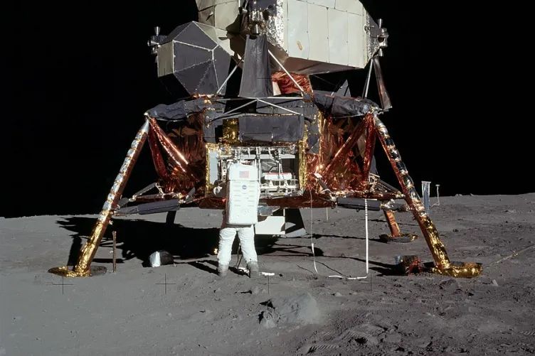 A lunar lander sits on the moon. A person in a space suit has their back to the camera, their arms on the lander.