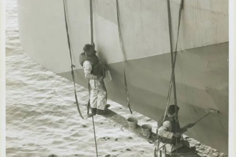 Two men are hanging by ropes from the side of a ship. They have paint cans and paint rollers.