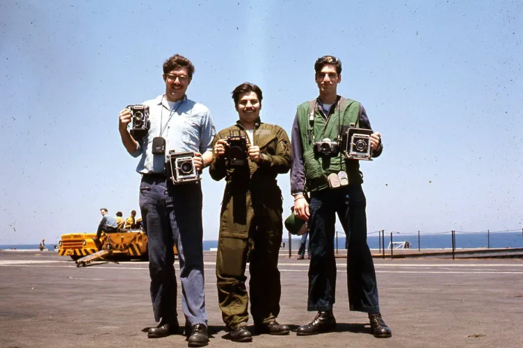 Archival photograph of three men standing on the flight deck holding cameras.