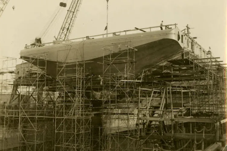Archival photograph of many scaffolding structures holding up a ship. People are standing on various parts of the scaffolding. A crane is in the background.