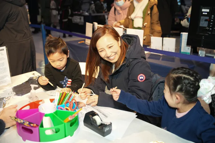 A woman engages with two young children in a creative activity involving drawing and crafts.