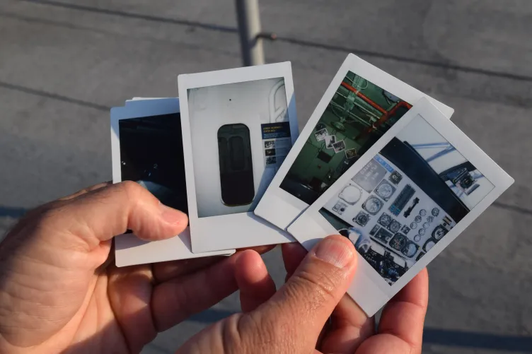 A hand holds a stack of Polaroid photos showcasing various interior views of The Intrepid, including control panels and operational spaces.