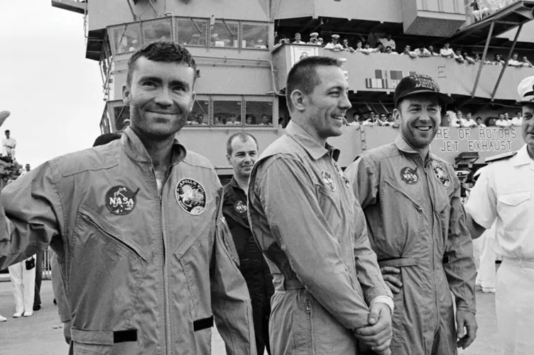 Four astronauts in flight suits, one waving, are greeted by a naval officer on the deck of an aircraft carrier, celebrated by onlookers for their achievements in space exploration.