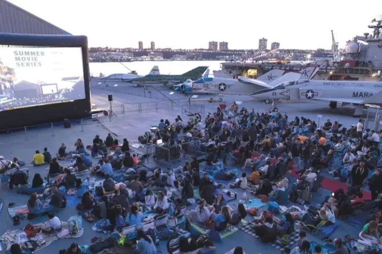 A large group of people are watching a movie on the flight deck of an aircraft carrier, surrounded by planes.