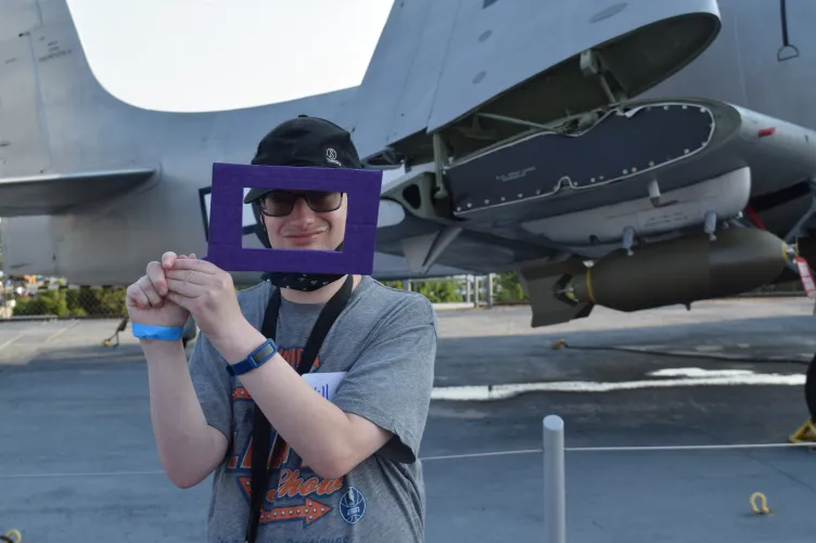 A young man wearing glasses and a black cap holds up a purple rectangular frame in front of an airplane.