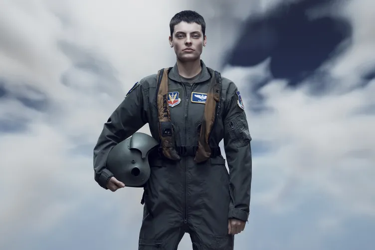 A person in a military flight suit, holding a helmet, stands confidently against a dramatic, cloudy sky.