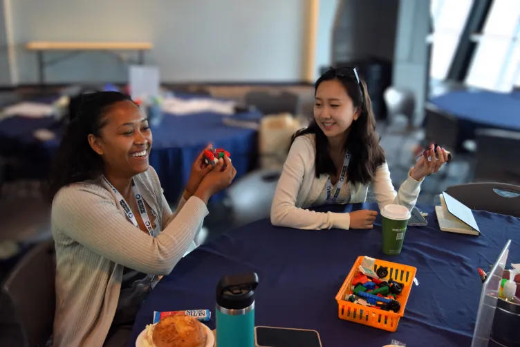 Two women seated at a round table at a conference in the Intrepid Education Center, closely examining and inspecting objects in their hands.