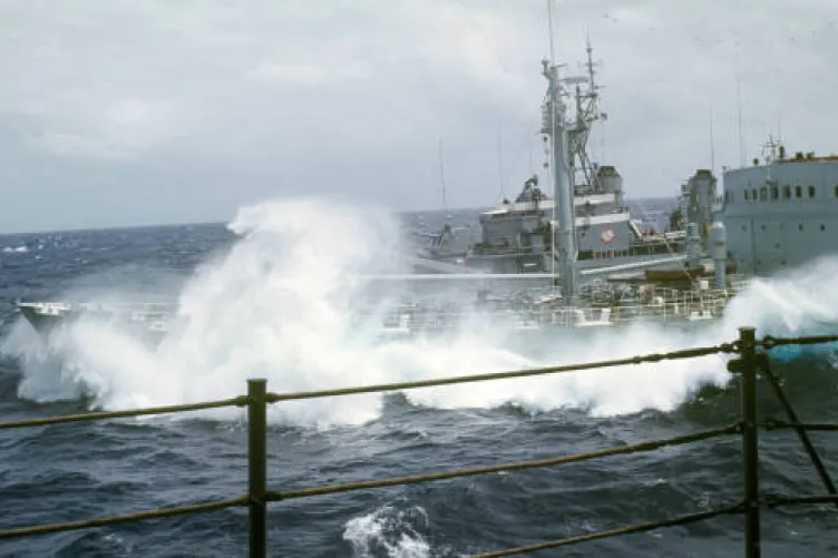 Archival image of water crashing over the bow of a ship that is pulling alongside Intrepid. The railing of Intrepid is in the foreground. The water is choppy and the sky is gray and cloudy.