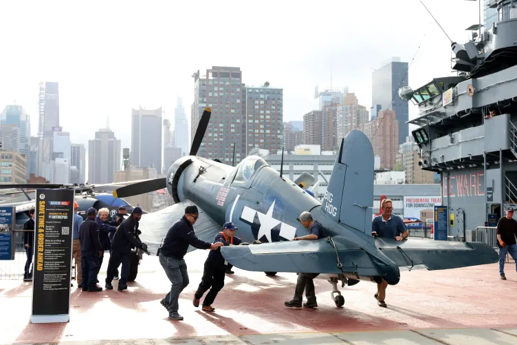 orsair aircraft on the flight deck at the Intrepid Museum, with a group of men pushing it toward the aircraft restoration tent, and the NYC skyline in the background.