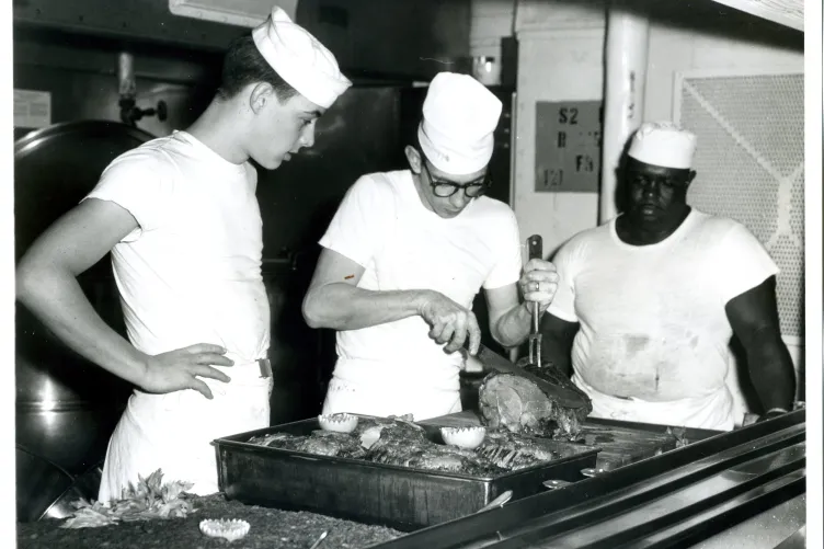 Sailors wearing chef hats carving a piece of meat in the kitchen aboard the Intrepid Museum.