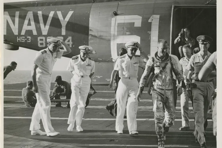 Astronaut Scott Carpenter walking out of a helicopter on a flight deck, surrounded by Naval officers.