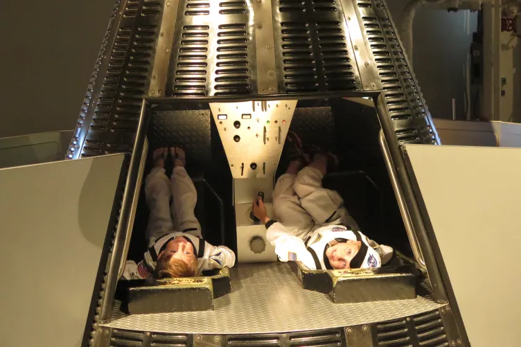 Two children in space suit costumes, sitting in a replica Gemini capsule.