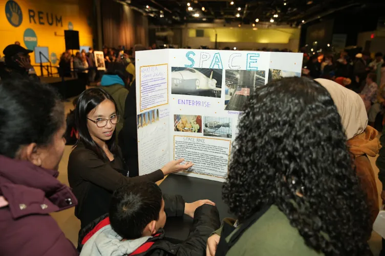 A young woman presenting a science project to several onlookers