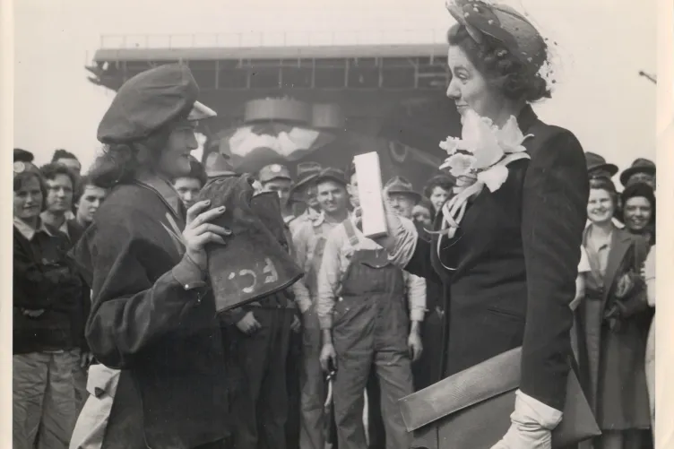 Black and white photograph showing a group of people, with two women prominently in the foreground engaged. One woman wears a uniform and cap, while the other wears a formal outfit with gloves, a large flower corsage, and a decorative hat.